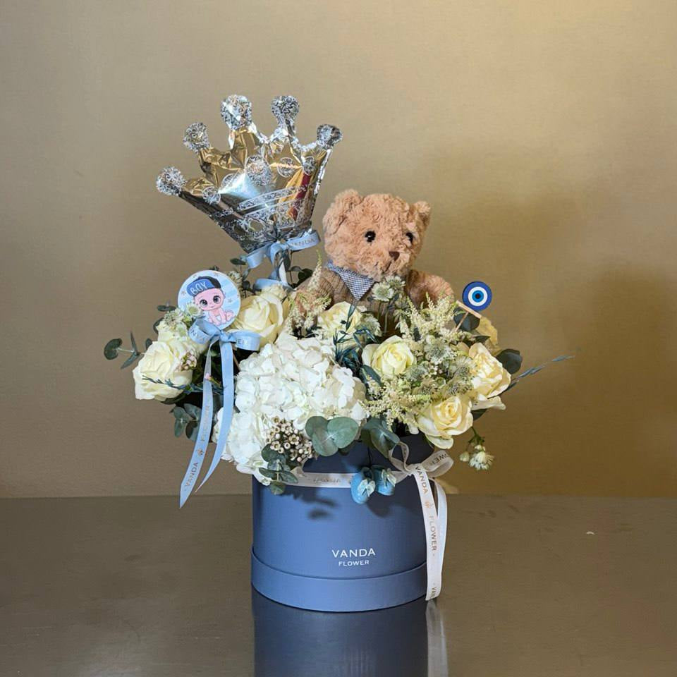 Baby boy themed flower arrangement in a blue round box with white roses, hydrangea, teddy bear, silver crown balloon, and decorative ribbons.