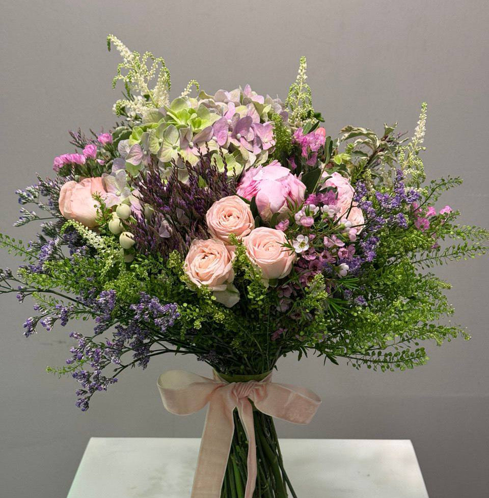 Pastel hand-tied bouquet with pink roses, hydrangea, lilac flowers, and greenery tied with a velvet ribbon.