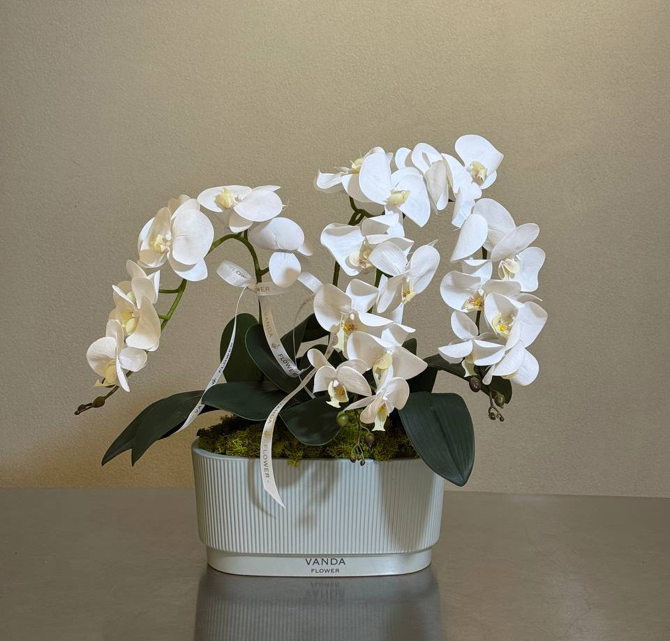 Artificial white Phalaenopsis orchid arrangement in a ribbed white ceramic planter.
