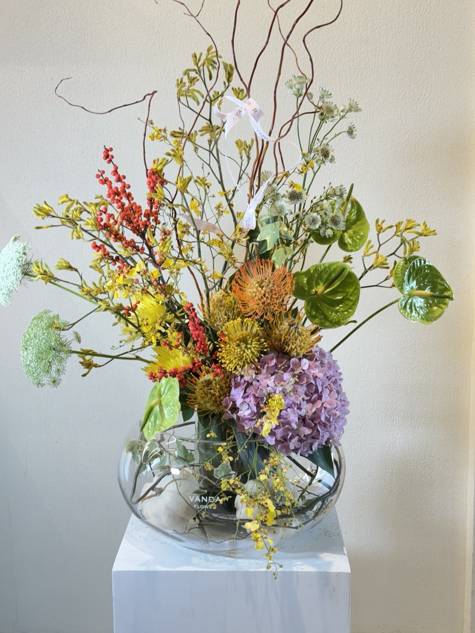A luxurious artistic arrangement featuring exotic flowers such as hydrangeas, proteas, anthuriums, and orchids beautifully styled in a clear glass bowl vase. A striking composition of texture, color, and modern design, perfect for elegant interiors or sophisticated gifting.