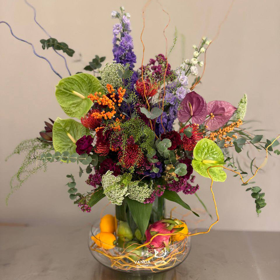Colorful fresh flower arrangement with anthurium, chrysanthemums, berries, and garden blooms in a glass bowl with citrus accents.
