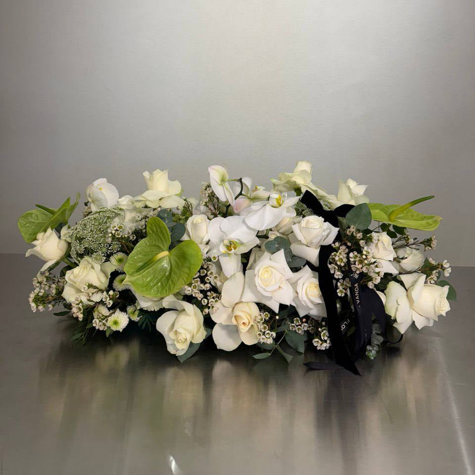 Long low white sympathy centerpiece with white roses, phalaenopsis orchids, green anthurium, waxflower, Queen Anne’s lace, and eucalyptus with black ribbon