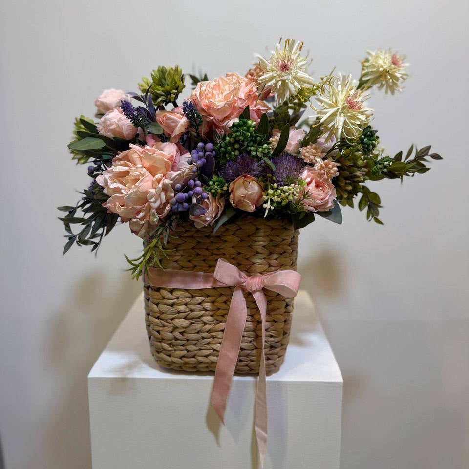 A beautifully arranged wicker flower basket filled with pink, purple, and white flowers, tied with a soft ribbon, perfect for gifting on special occasions