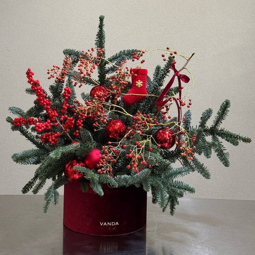 
Christmas-themed floral arrangement with red ornaments, berries, and evergreen branches in a velvet red box.